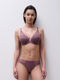 A woman modeling a matching dusty rose purple lingerie set consisting of a seamless lace bra and coordinating panties, standing against a plain light gray wall.
