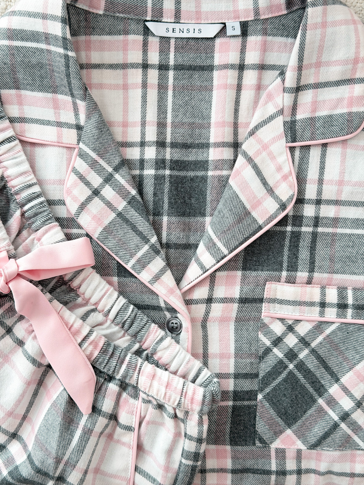 A close-up of a plaid pajama set with a pink bow, featuring a button-front top and matching pants in gray, pink, and white plaid with pink piping. The brand name 'SENSIS' is visible on the inner neck label.
