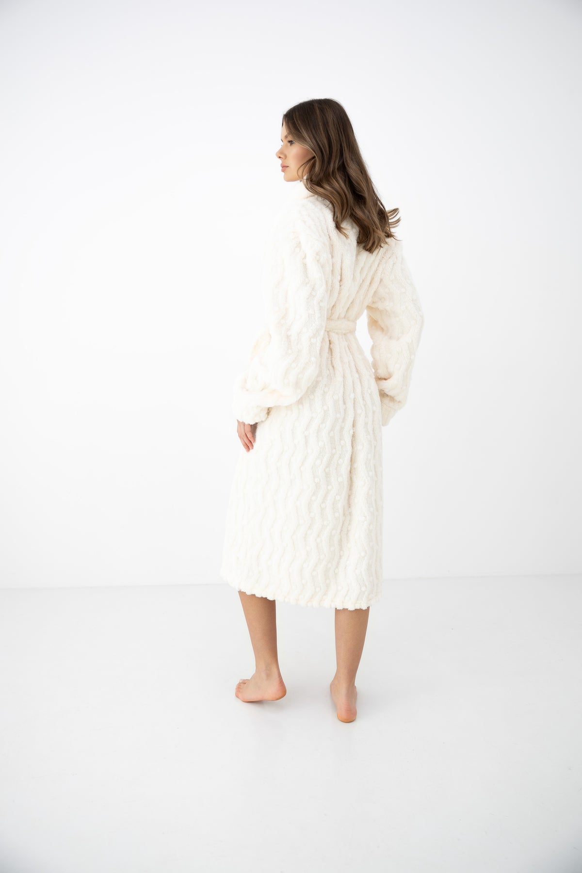 A woman wearing a cream-colored, textured knit robe is shown from the back in a studio setting with a white background.