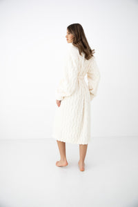 A woman wearing a cream-colored, textured knit robe is shown from the back in a studio setting with a white background.