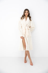 A woman modeling a plush, textured cream-colored bathrobe with a tie belt, standing against a seamless white studio background.