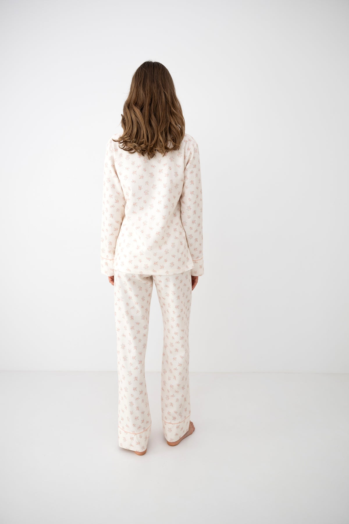 A woman viewed from behind wearing a matching long-sleeve pajama top and pants set in a warm off-white fabric with a small beige floral pattern, set against a plain white studio background.