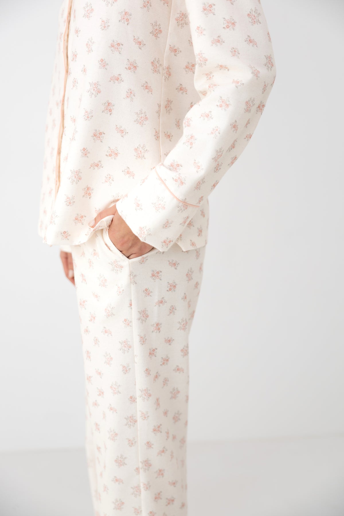 A person wearing a matching long-sleeve button-front top and pants pajama set in a warm off-white fabric with a small repeating pink floral print, shown against a plain light background.