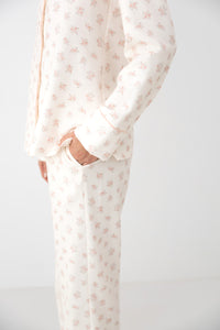 A person wearing a matching long-sleeve button-front top and pants pajama set in a warm off-white fabric with a small repeating pink floral print, shown against a plain light background.