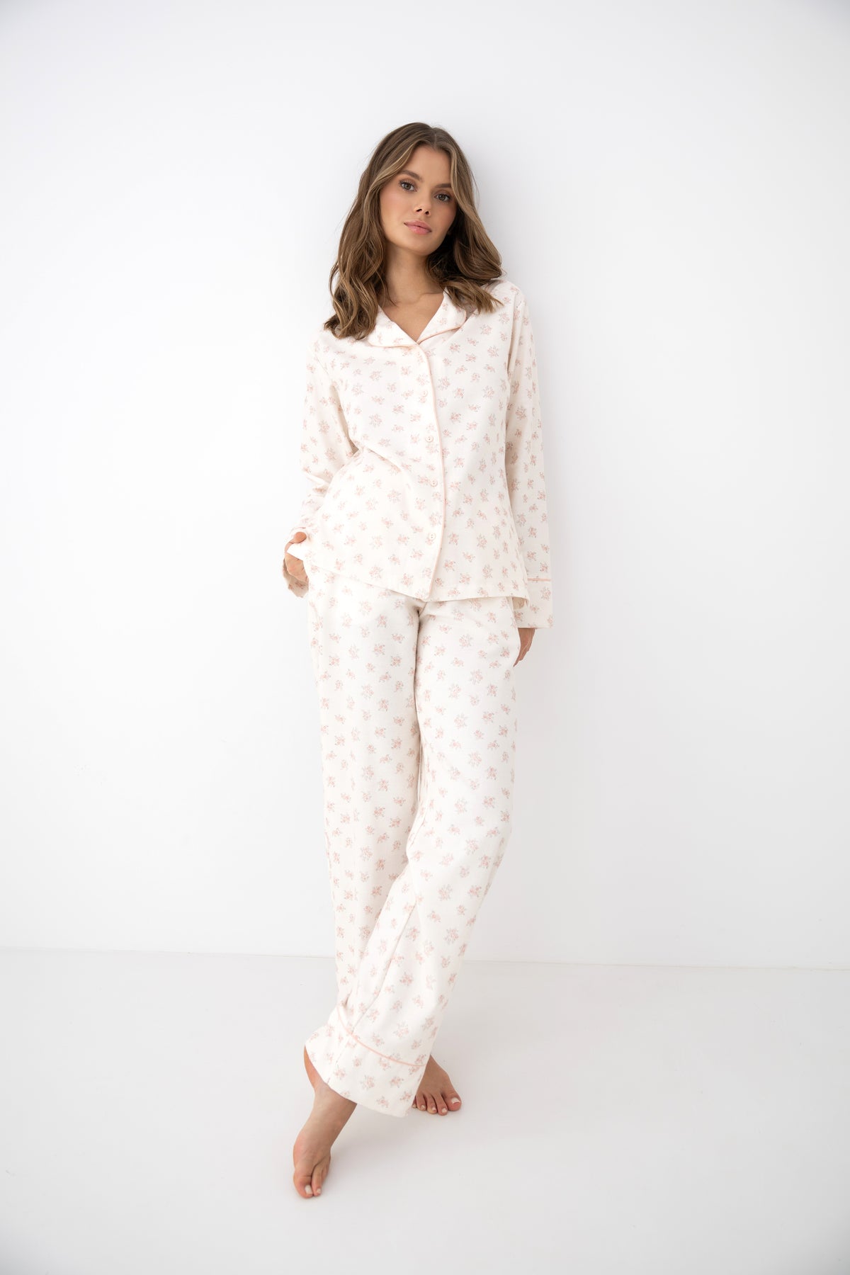 A woman modeling a matching two-piece pajama set with a long-sleeve button-up top and relaxed pants, featuring a soft cream base with a delicate light pink floral pattern, set against a plain white studio background.