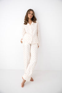 A woman modeling a matching two-piece pajama set with a long-sleeve button-up top and relaxed pants, featuring a soft cream base with a delicate light pink floral pattern, set against a plain white studio background.