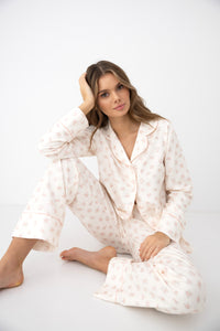 A woman wearing a light cream pajama set with a delicate pink floral pattern, sitting on a white studio floor.