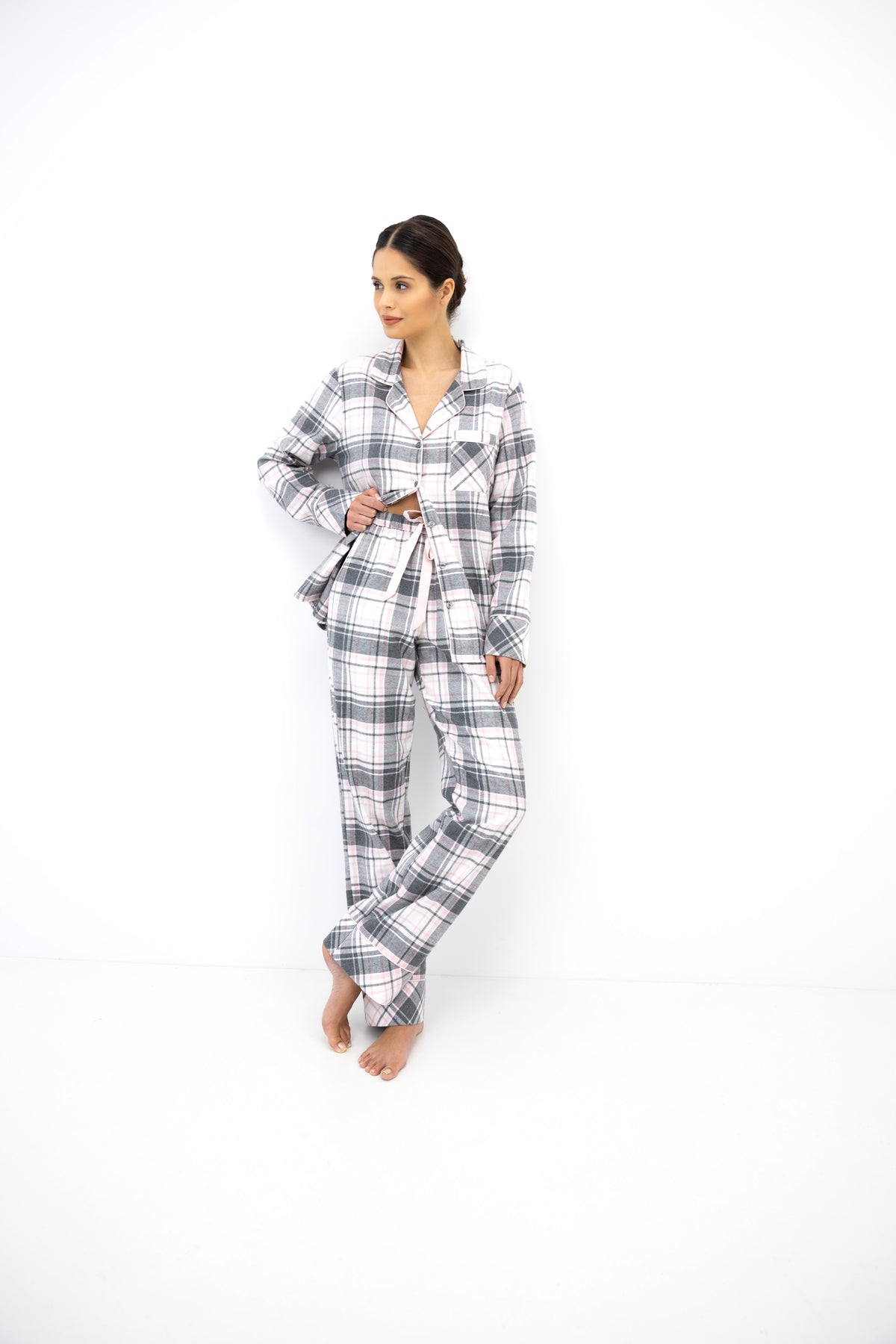 A woman modeling a matching plaid pajama set consisting of a long-sleeve button-up shirt and wide-leg pants, standing against a plain white studio background.