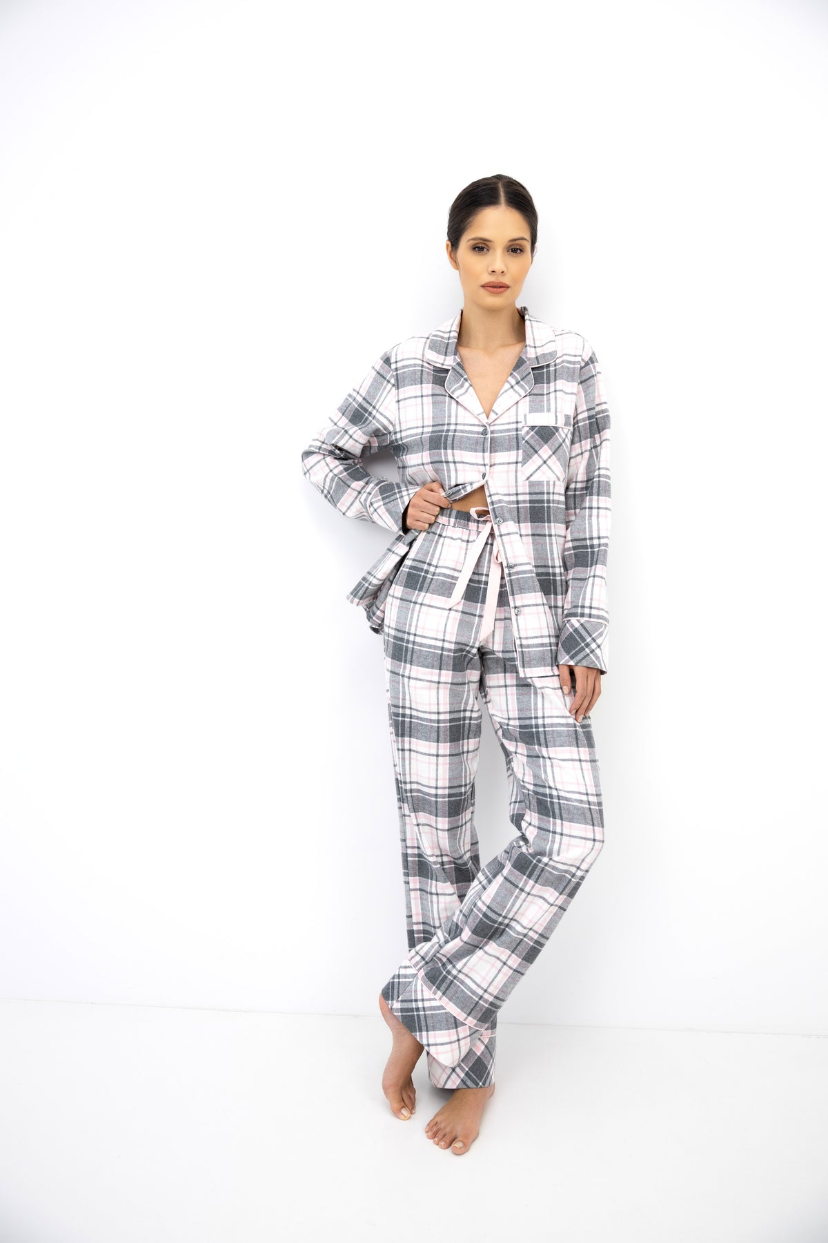 A woman modeling a matching plaid pajama set consisting of a long-sleeve button-up shirt and wide-leg pants, featuring a gray, white, and black checkered pattern, against a plain white studio background.