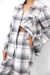 A model wearing a matching plaid pajama set with a long-sleeve button-front top and coordinating pants in a gray, white, and light pink plaid pattern.