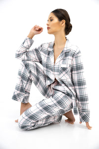 A woman kneeling on a white studio floor wearing a matching plaid pajama set with a long-sleeve button-front top and pants. The fabric has a white base with gray and light pink plaid lines.