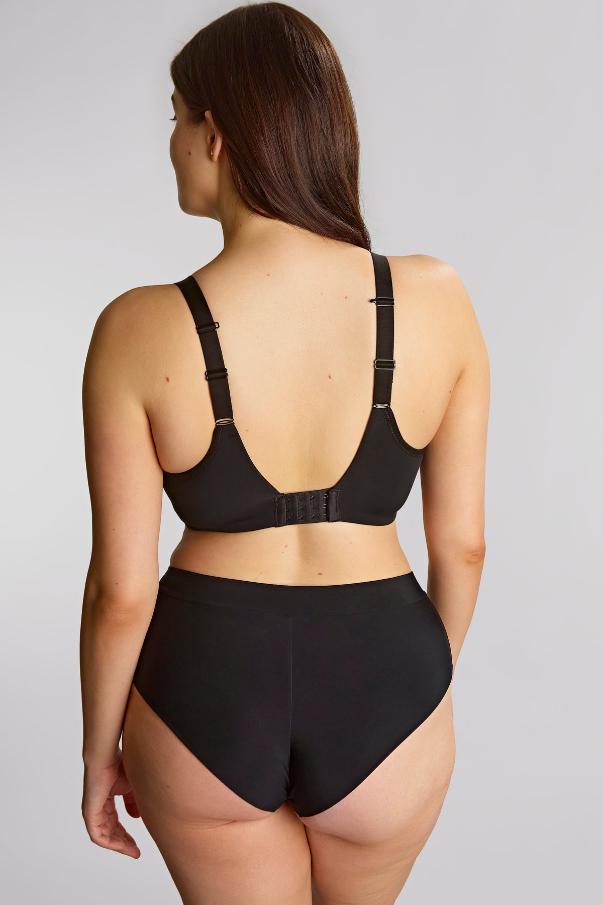 A woman shown from the back wearing a black bralette with adjustable shoulder straps and matching high-waisted briefs against a light gray studio background.