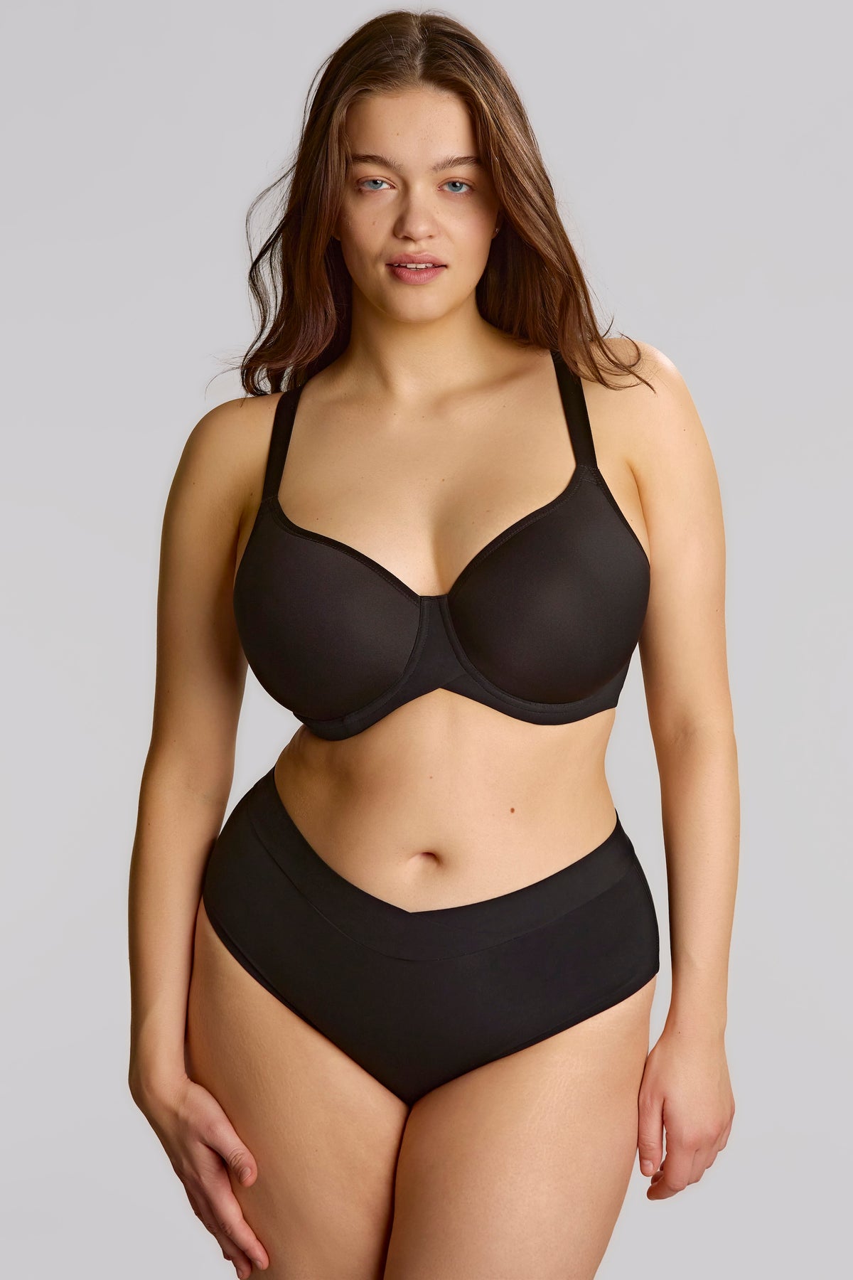 A woman modeling a black two-piece lingerie set consisting of a seamless underwire bra and matching high-waisted briefs, shot against a solid gray studio background.