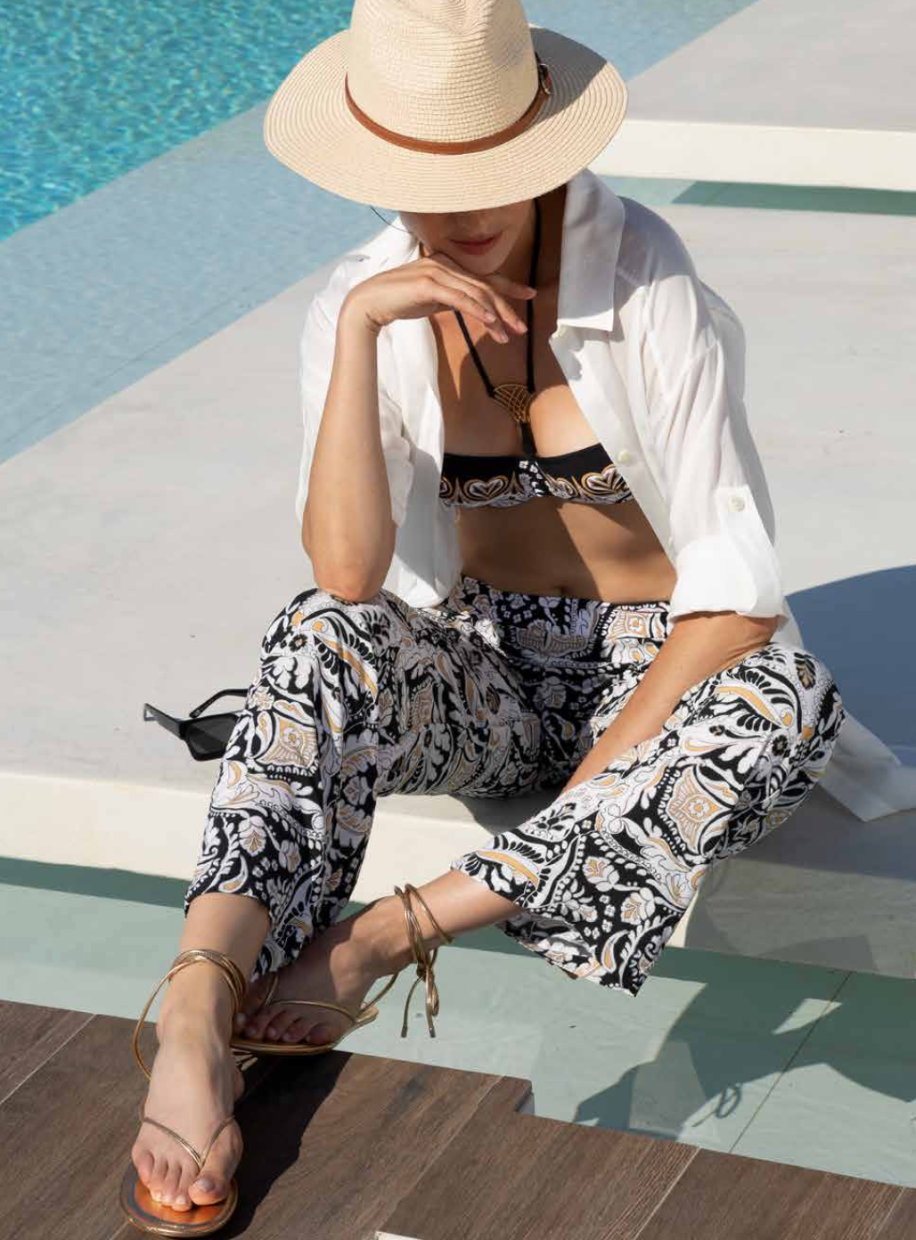 Woman in a white shirt and patterned pants sitting by a pool.