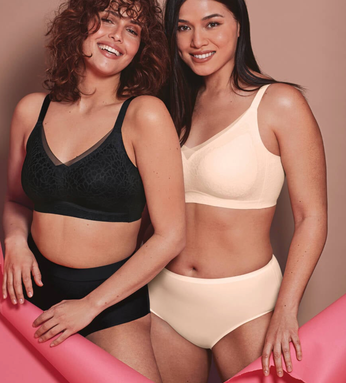 Two women wearing black and beige bras and panties on a pink surface.