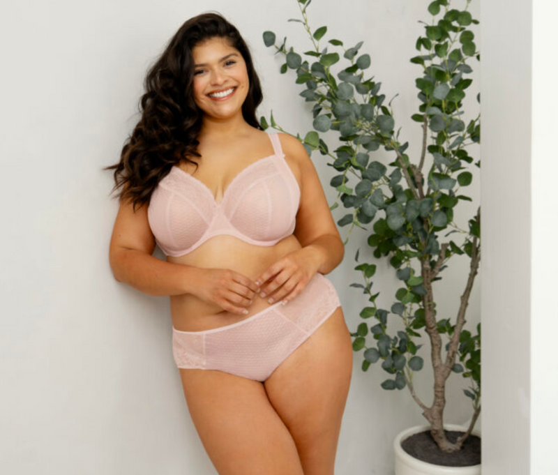 Woman wearing a pink bra and underwear set standing next to a plant against a white wall.