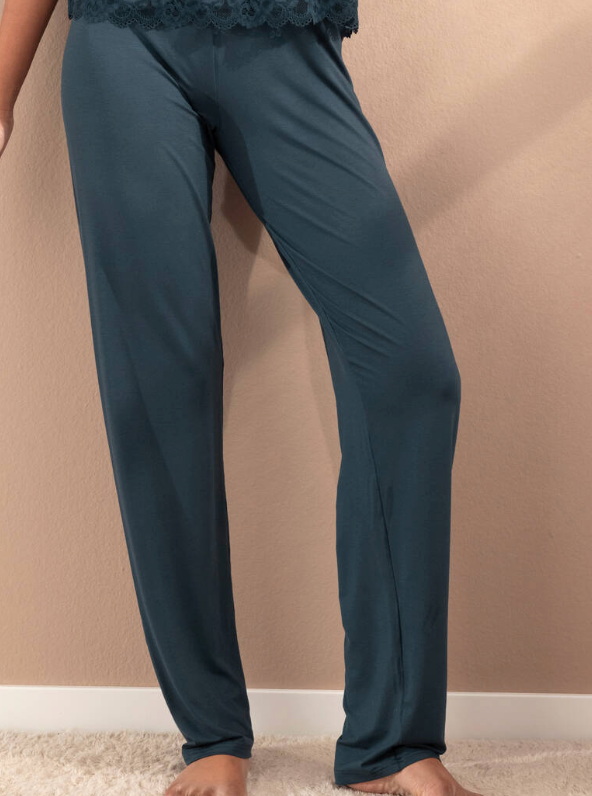 A person wearing loose, straight-leg pajama pants in a muted blue-green/teal color, standing barefoot on a light carpet against a beige wall.