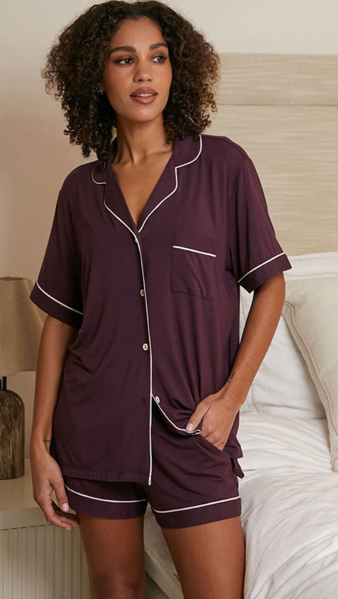 A woman modeling a two-piece pajama set consisting of a short-sleeve button-front top and matching shorts in deep plum purple with thin white piping trim, set against a bedroom background with a bed and lamp.