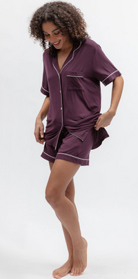 A woman modeling a matching short-sleeve button-front pajama shirt and shorts set in deep plum purple with thin white piping trim, photographed against a clean white studio background.