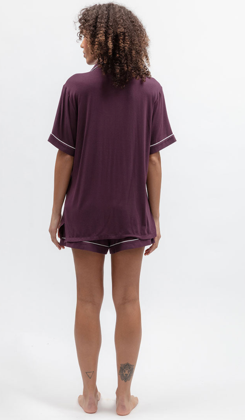A woman is shown from behind wearing a matching short-sleeve t-shirt and shorts pajama set in a deep plum purple color, featuring thin white piping details on the sleeve cuffs and hem. She is standing barefoot against a clean white studio background.