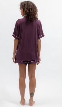 A woman is shown from behind wearing a matching short-sleeve t-shirt and shorts pajama set in a deep plum purple color, featuring thin white piping details on the sleeve cuffs and hem. She is standing barefoot against a clean white studio background.