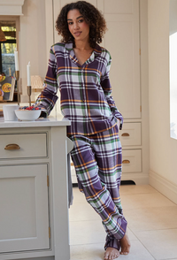 A woman wearing a purple, green, and white plaid pajama set stands barefoot in a bright kitchen, leaning against a white cabinet next to a counter with mugs and a bowl.