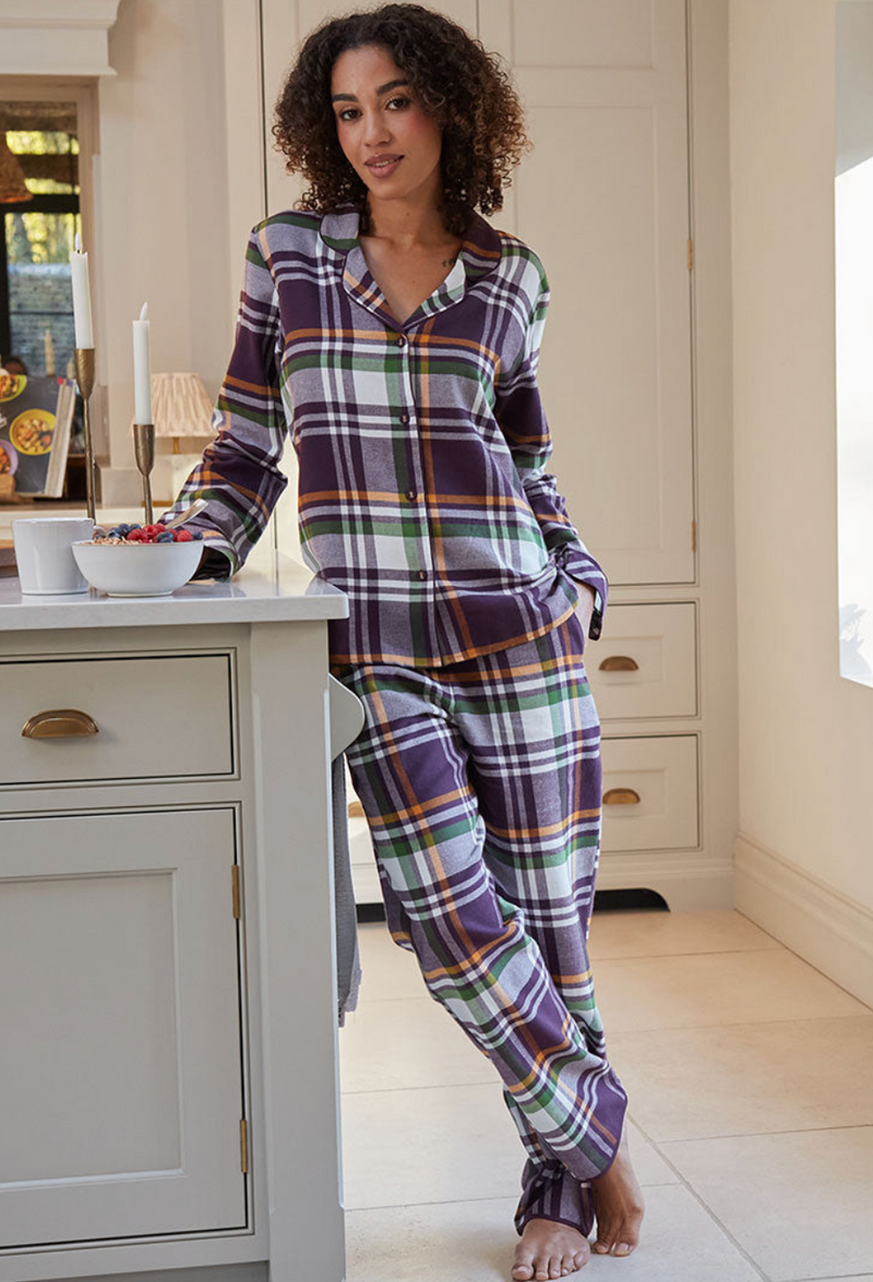 A woman wearing a purple, green, and white plaid pajama set stands barefoot in a bright kitchen, leaning against a white cabinet next to a counter with mugs and a bowl.