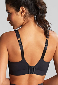 Rear view of a woman wearing a black wireless sports bra with adjustable underband straps and a center closure, shown against a light gray studio background.
