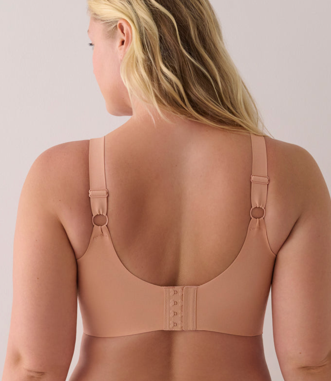 A woman shown from the back wearing a seamless, light tan/nude bra with adjustable straps and a hook-and-eye closure.