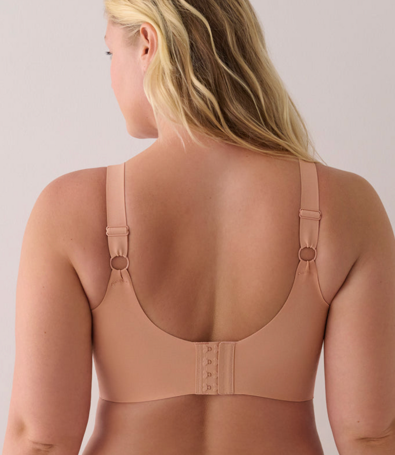 A woman shown from the back wearing a seamless, light tan/nude bra with adjustable straps and a hook-and-eye closure.