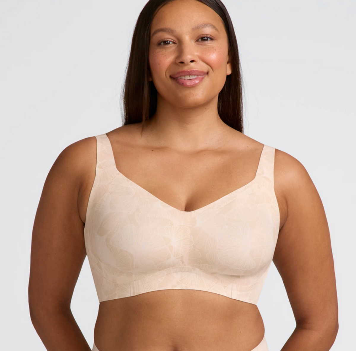 A woman modeling a light beige underwire bra with a subtle tonal floral pattern, shown against a solid light gray background.