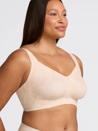A close-up of a woman wearing a light beige seamless bra with a subtle floral jacquard pattern, shown from the chest up against a solid light gray background.