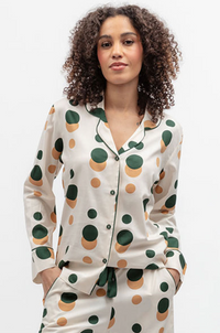 A woman modeling a matching pajama set with a long-sleeve button-front top and coordinating pants. The pajamas have a pink base with large abstract polka dots in dark green and tan, with dark green piping.