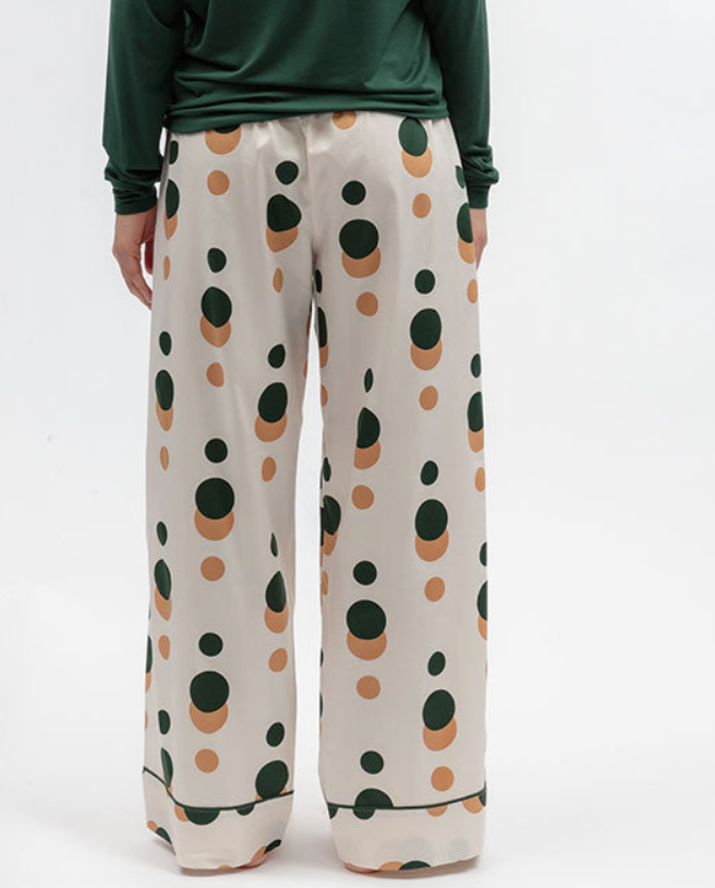 A person wearing wide-leg pajama pants with a pink cream base and dark green and tan polka dots, paired with a dark green long-sleeve top, shown from the waist down on a white studio background.