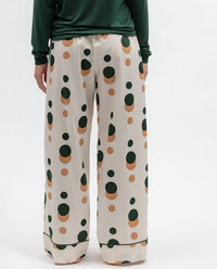 A person wearing wide-leg pajama pants with a pink cream base and dark green and tan polka dots, paired with a dark green long-sleeve top, shown from the waist down on a white studio background.