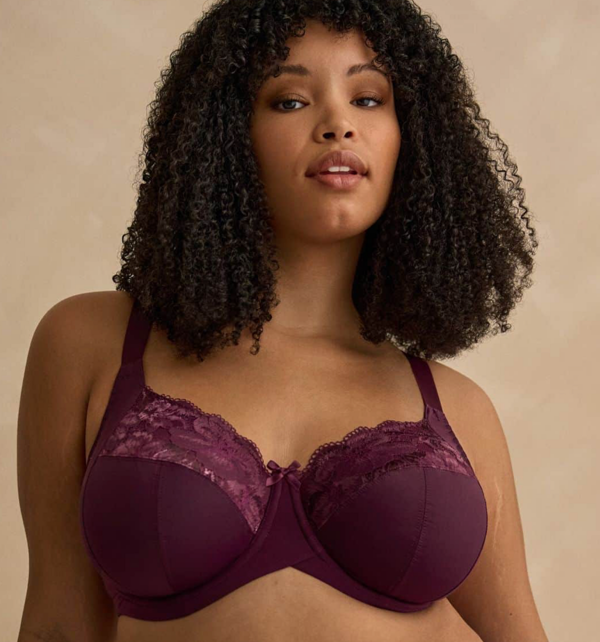 A woman with voluminous dark curly hair wears a deep plum purple underwire bra with lace cups, photographed against a warm beige studio background.