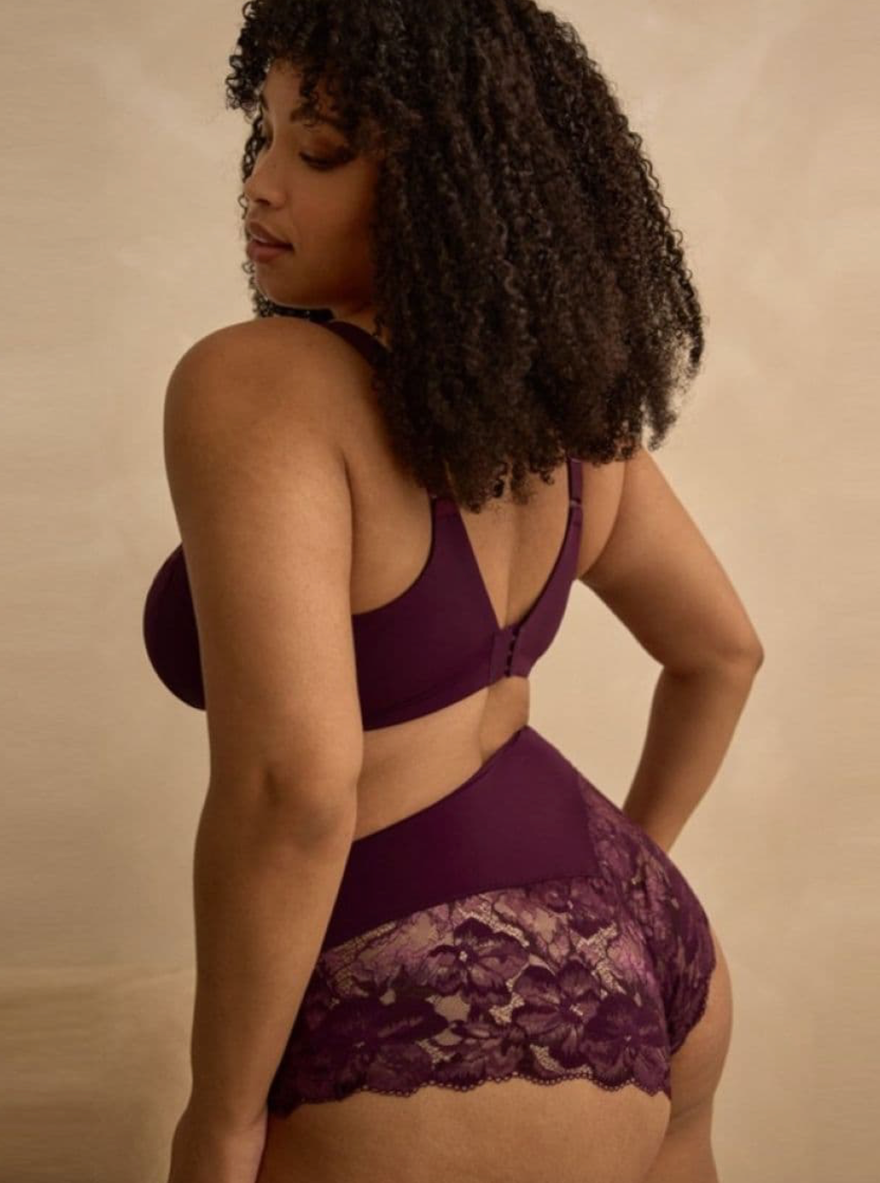 A woman with curly dark hair is shown from the back wearing a deep purple lingerie set with a lace-trimmed bottom and a matching bra, posed against a warm beige studio backdrop.