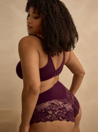 A woman with curly dark hair is shown from the back wearing a deep purple lingerie set with a lace-trimmed bottom and a matching bra, posed against a warm beige studio backdrop.