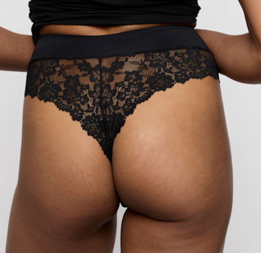 Rear view of a person wearing black lace thong underwear against a light gray studio background.