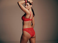 A woman posing from the back wearing a red lace bra and matching red lace underwear against a plain warm gray studio background.