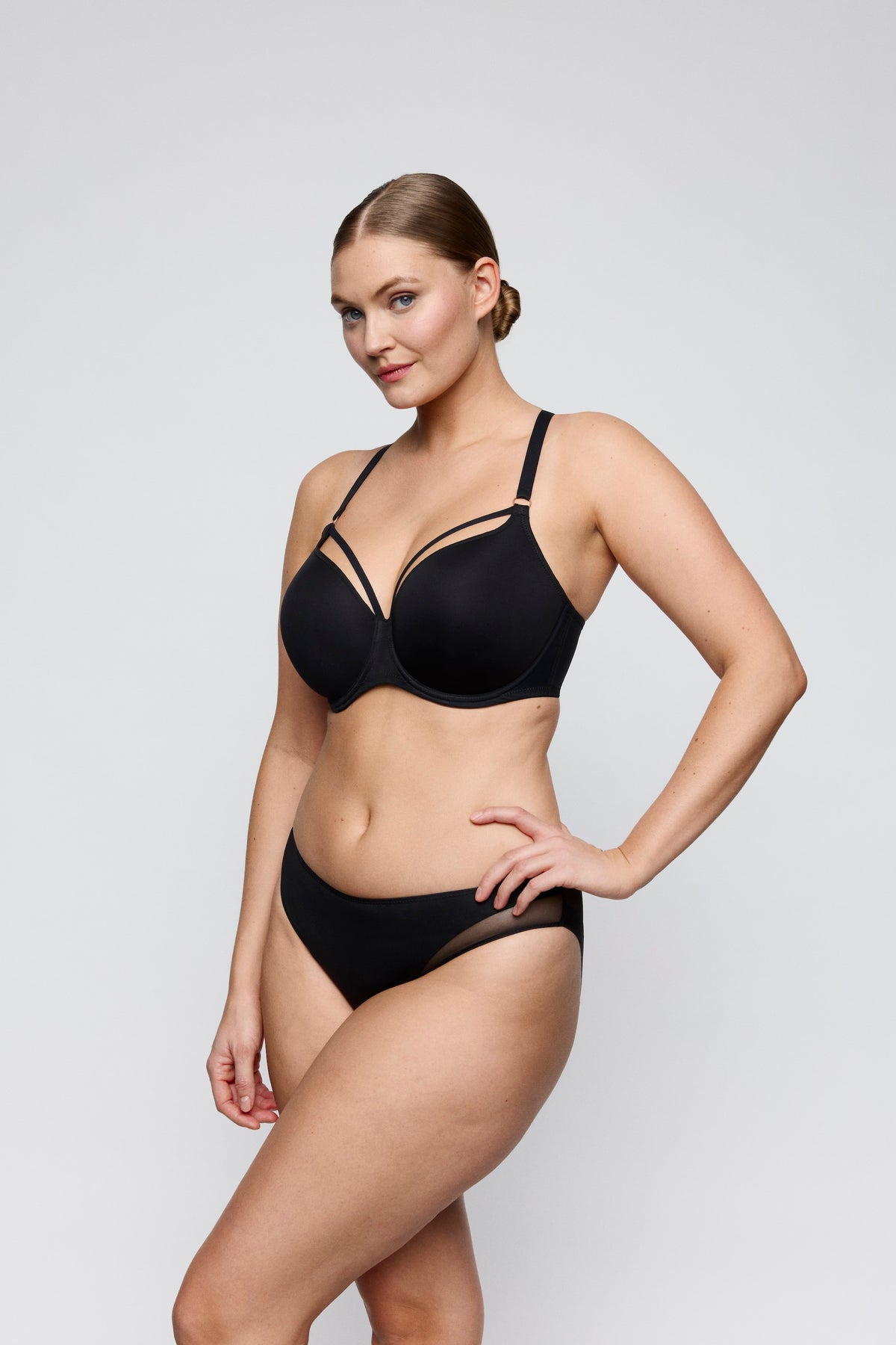A woman modeling a black two-piece lingerie set consisting of a strappy underwire bra and matching high-waisted briefs, photographed against a light gray studio background.