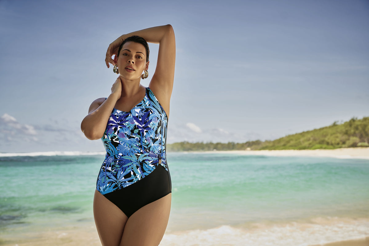 Woman in a blue and black swimsuit standing on a beach with clear blue water and greenery in the background.