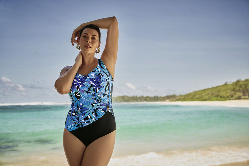 Woman in a blue and black swimsuit standing on a beach with clear blue water and greenery in the background.